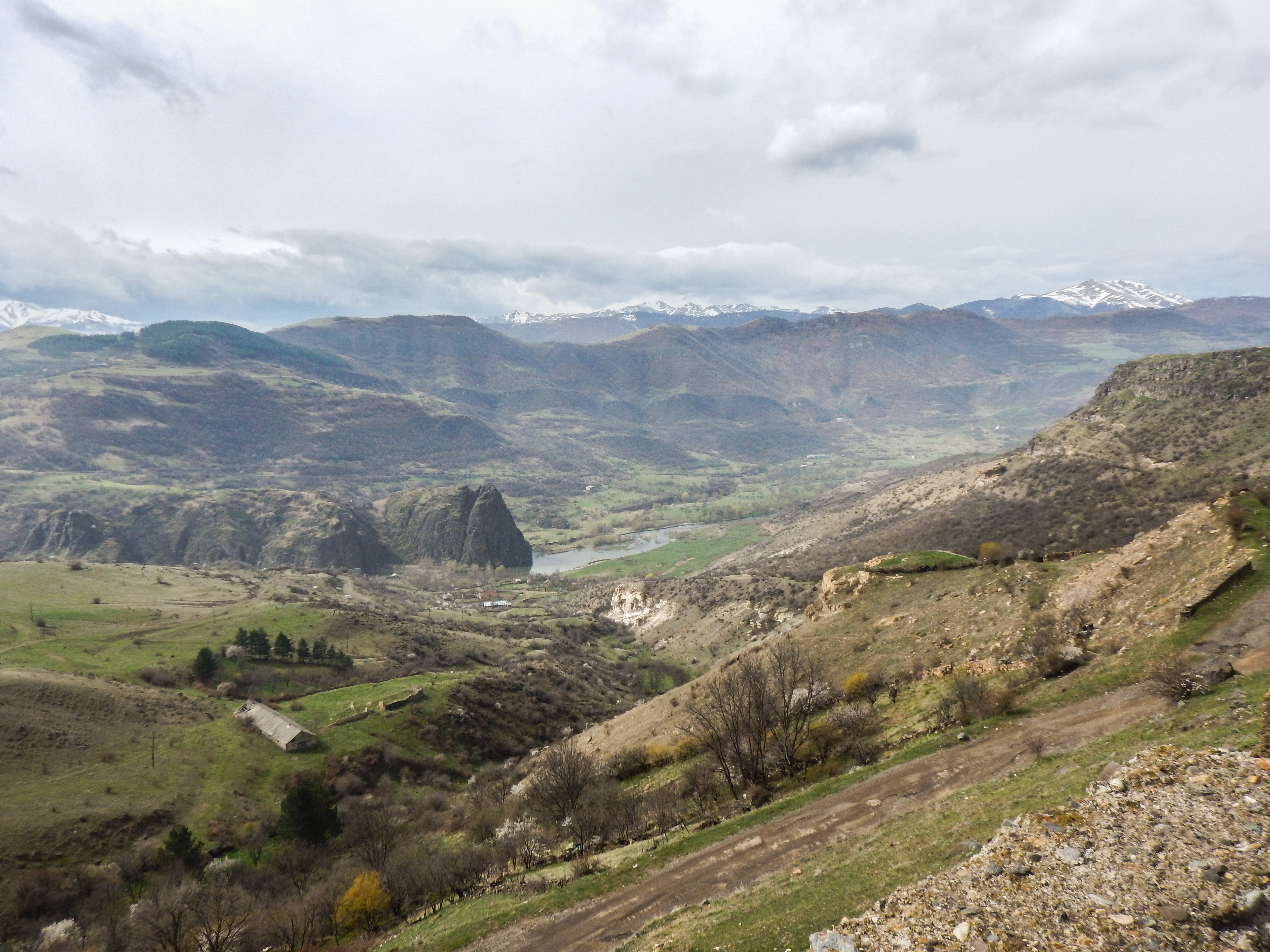 A mountain valley landscape in the Southern Caucasus with grassy hills, scattered trees, rocky slopes, and a river running through the centre; snow-capped peaks visible in the distance under a cloudy sky.