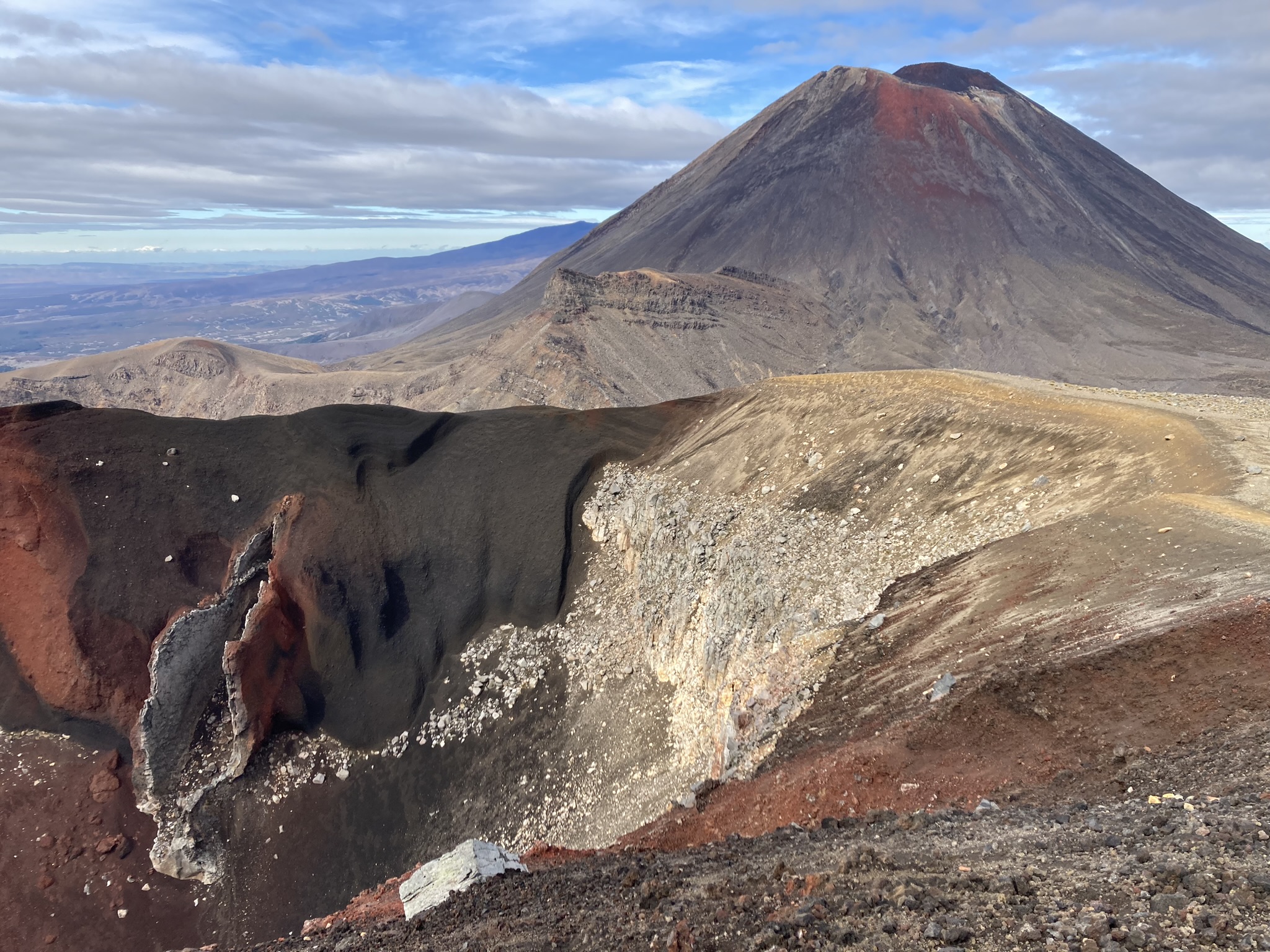 This photograph shows Ngāuruhoe in the background – a cone shaped volcano that is mostly black and grey, with red lavas at the top that last erupted in 1975. In the foreground is vertical fissure, that is part of Red Crater, that last erupted in the 1920’s. Black, red, and yellowy-brown volcanic rocks such as scoria and lava flows can be seen throughout.