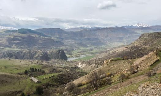 A mountain valley landscape in the Southern Caucasus with grassy hills, scattered trees, rocky slopes, and a river running through the centre; snow-capped peaks visible in the distance under a cloudy sky.