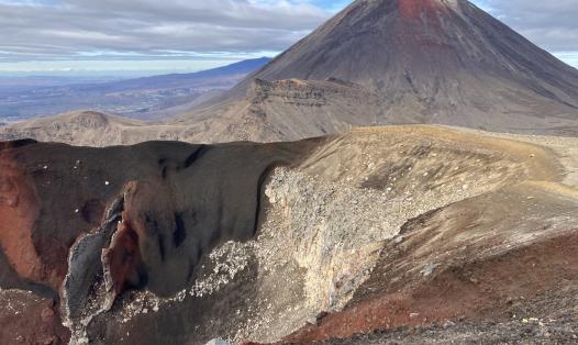 This photograph shows Ngāuruhoe in the background – a cone shaped volcano that is mostly black and grey, with red lavas at the top that last erupted in 1975. In the foreground is vertical fissure, that is part of Red Crater, that last erupted in the 1920’s. Black, red, and yellowy-brown volcanic rocks such as scoria and lava flows can be seen throughout.