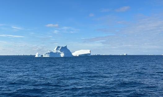 A large white iceberg on the horizon under a bright sky with deep blue ocean water and a few light clouds.