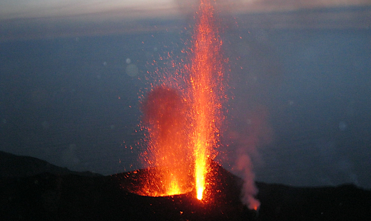 Image of a lava fountain explosion at the summit of Stromboli volcano. Image taken in the evening with the dark sea in the background. Foreground is of a hot yellow to red vertical fountain of lava erupting from two vents within a dark area at the volcano summit. A third vent to the right has noticeable grey outgassing which is partly illuminated red.  